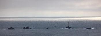 Longships Lighthouse long exposure This landscape photograph shows Longships Lighthouse positioned on a rocky outcrop in the sea off the coast of Cornwall. The image was captured on a late afternoon in early summer, with soft, diffused light illuminating the expanse of water and the surrounding rocks of the Longships reef. The lighthouse stands prominently as the main subject of the composition, with the sea stretching out to the horizon and faint landmasses visible in the distance. Subtle movement in the water suggests a long exposure technique, resulting in a smooth texture across the surface of the sea.